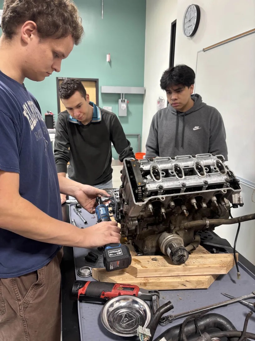 Three people looking at an engine sitting on a table.